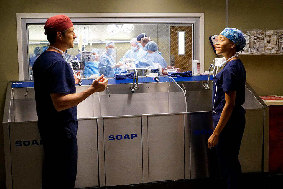 Two characters in medical scrubs stand in a hospital set, with a surgery in progress visible through a window behind them