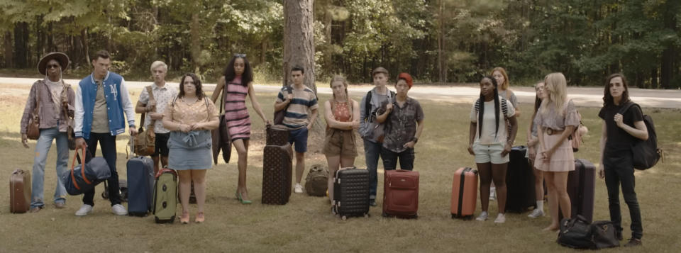 A group of young adults, each with a suitcase, stands in a grassy area surrounded by trees