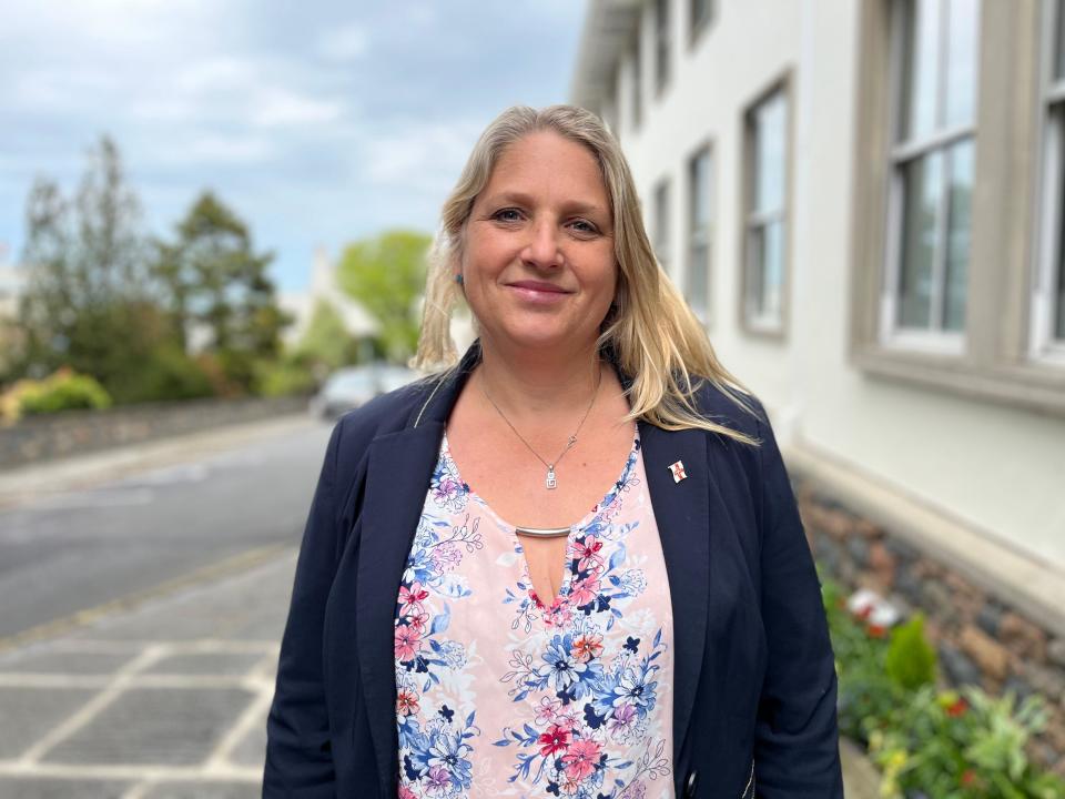 Deputy Victoria Oliver. A smiling blonde woman wearing a blue blazer and flowery blouse. She is standing on a street outside a modern cream building.