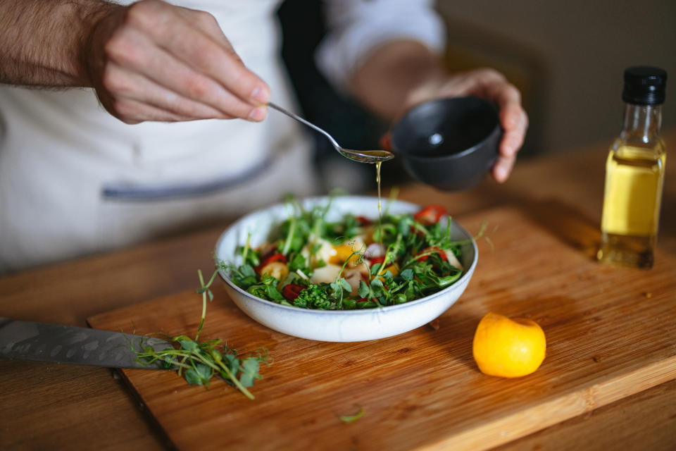 Close-up of a person drizzling dressing on a salad with various greens, tomatoes, and cheese on a wooden board with a lemon and a bottle of olive oil