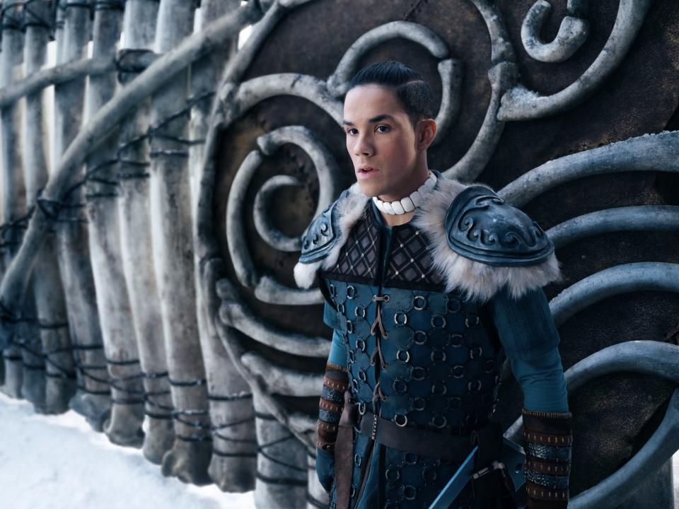 sokka in the live action avatar the last airbender, wearing blue, cold-weather battle gear and with his hair pulled back on top with an undercut. he's standing in front of a wall with a symbol of flowing waves on it