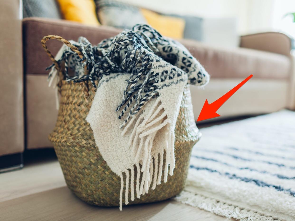 A basket with a blanket inside of it sits on the floor of a living room in front of a couch.