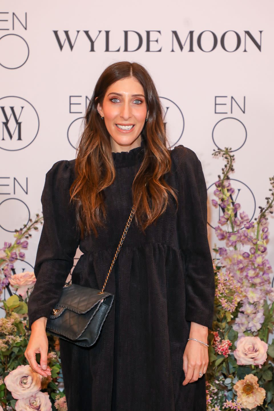 sarah jossel, a woman wearing a black long sleeve dress and cross body bag, long hair in loose waves, smiling at the camera