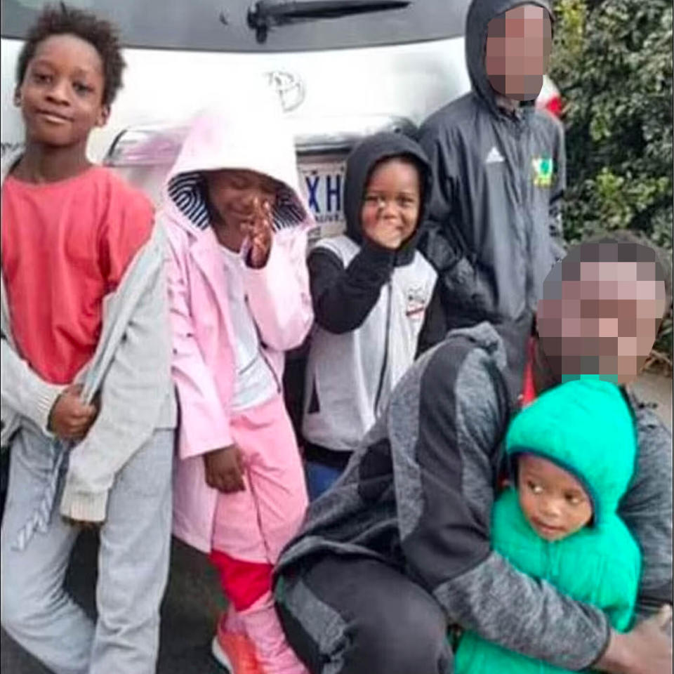 A photo showing four young children with tracksuits on smiling at  the camera. A man and younger child are standing to the right but with faces blurred.