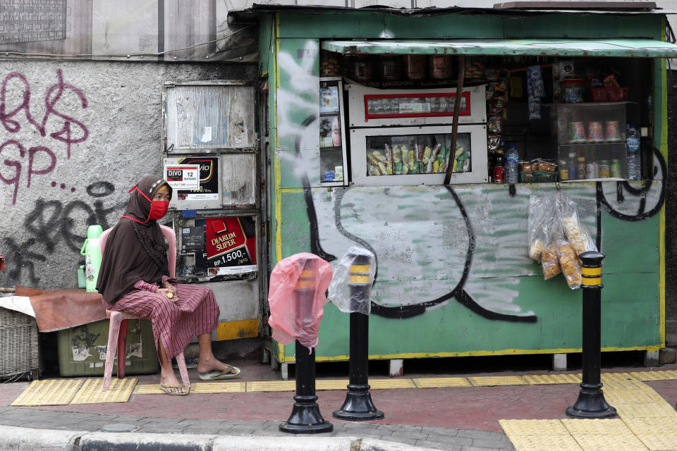 A woman wearing a face mask as a precaution against the coronavirus outbreak waits for customers near her roadside cigarette and snack stall in Jakarta, Indonesia, Friday, Aug. 14, 2020. An Indonesian vaccine production company has started this week a so-called phase 3 clinical trials to test a potential coronavirus vaccine developed by a Chinese company. (AP Photo/Tatan Syuflana)