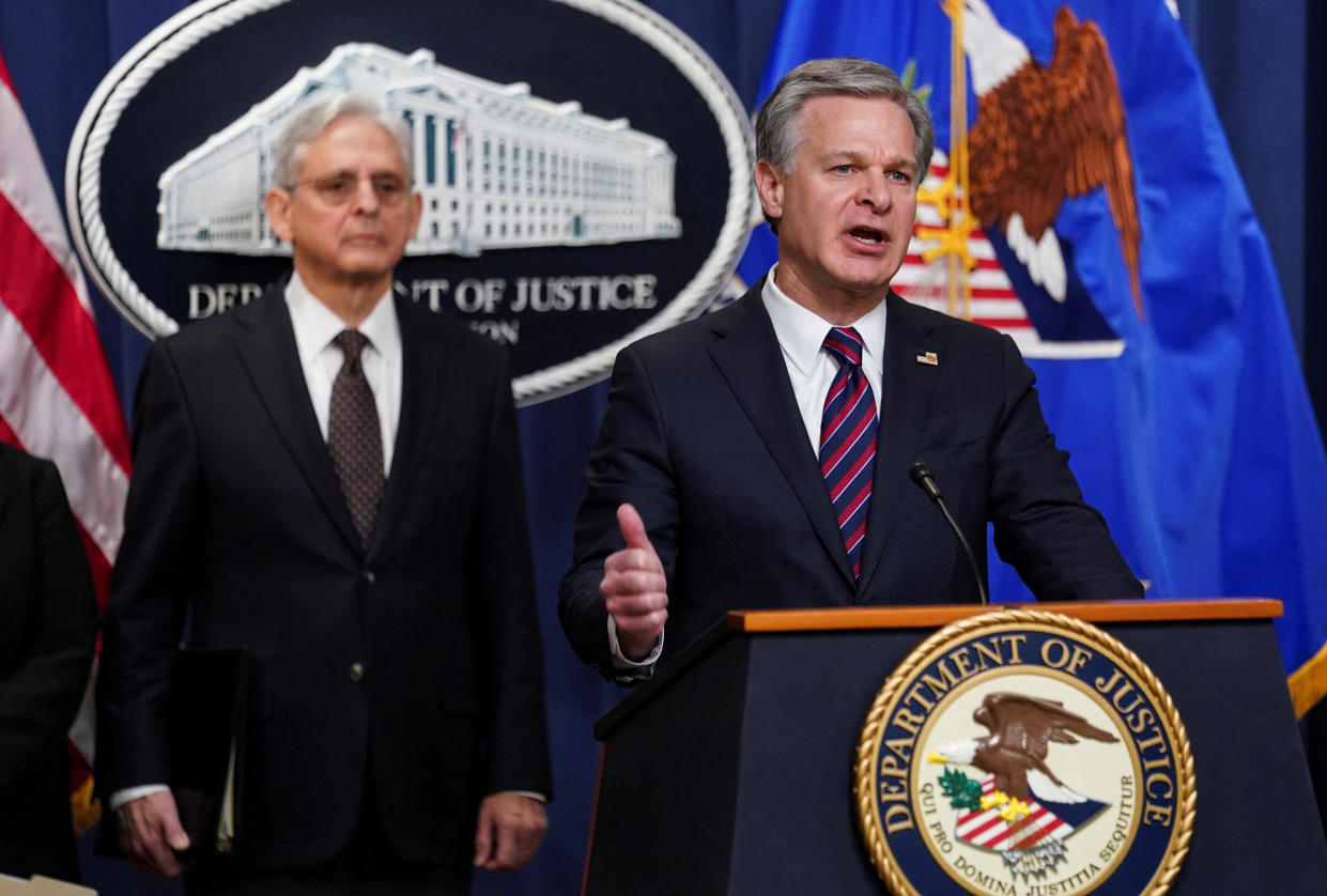 Christopher Wray at the podium, marked Department of Justice, with Merrick Garland standing behind him.
