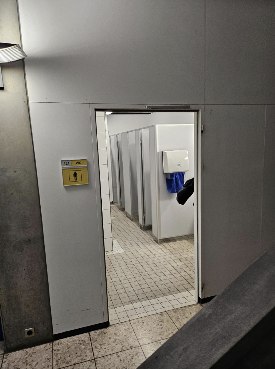 Public restroom interior with open stall doors and visible toilet paper dispenser; individual's foot seen under one door