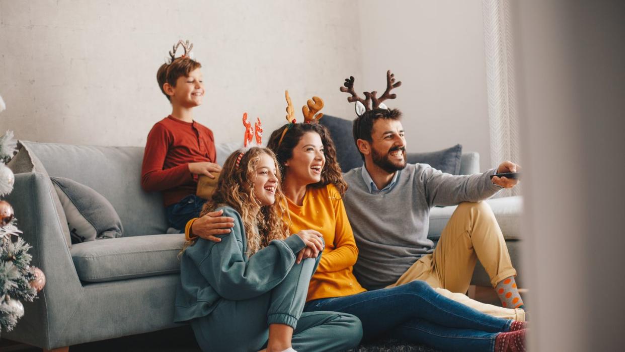 a young boy is sitting on the sofa bed, while his parents and his sister are on the floor they are all enjoying while they are watching tv christmas indoors