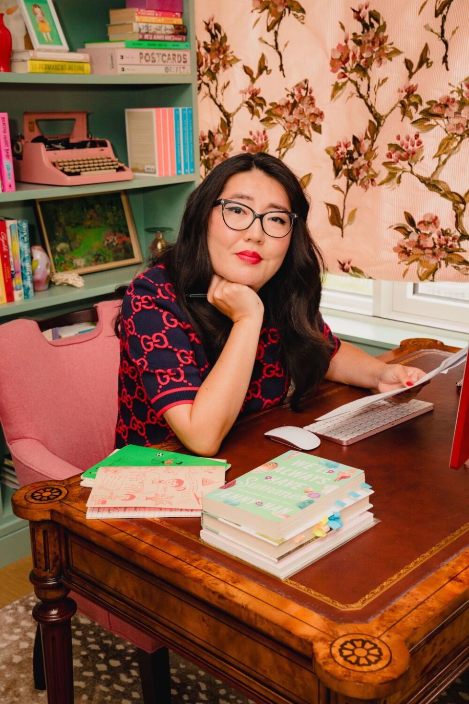 Jenny Han sits at a desk with stacked books and rests her hand under her chin.