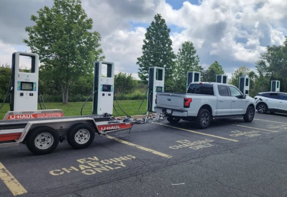 Several electric vehicle charging stations at a parking lot are being blocked by trucks towing trailers. The parking spots are marked "EV CHARGING ONLY."