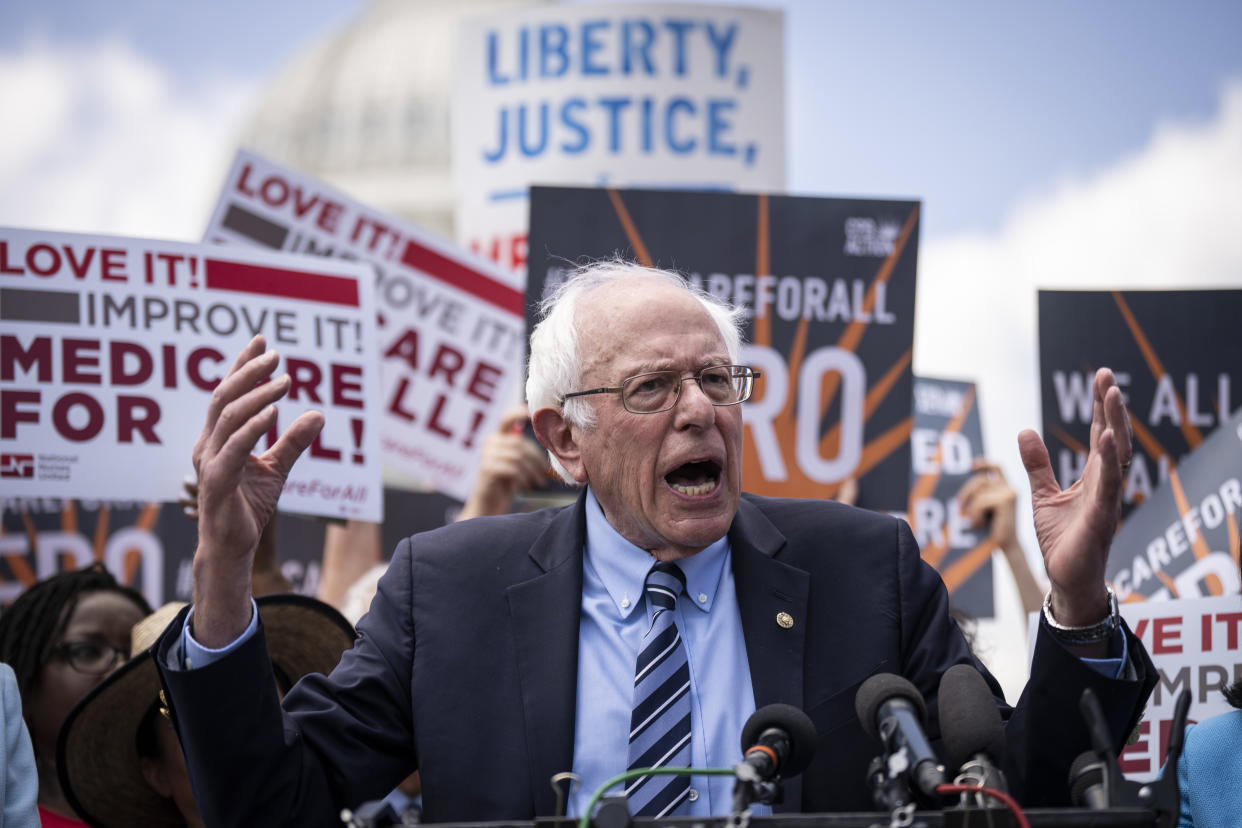 Sen. Bernie Sanders speaking during a news conference, with some people in the background holding signs calling for Medicare for All.