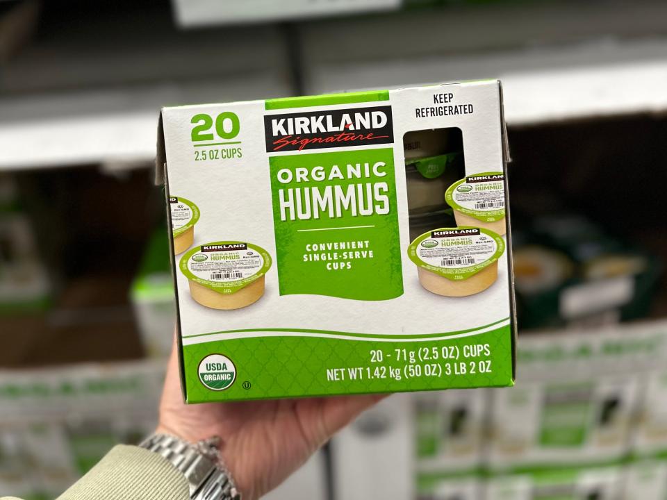 A hand holds a white and green box of organic hummus in front of boxes of guacamole and more containers of hummus at Costco