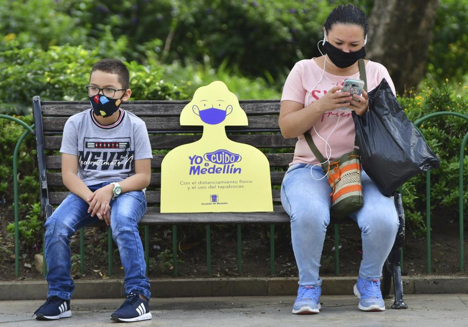 In this June 8, 2020 photo, a sign reminding citizens to maintain a safe social distance sits on a park bench between a woman and child, amid the new coronavirus pandemic in Medellin, Colombia, which recently went five weeks without a single COVID-19 death. The new challenge for Medellin will be to convince citizens to continue abiding by safety measures like wearing face masks and social distancing. In some poor neighborhoods, local activists say they've encountered skepticism about the virus. (AP Photo/Luis Benavides)