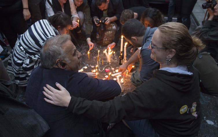 A man and woman sit arm in arm as others light candles on the ground at a vigil.