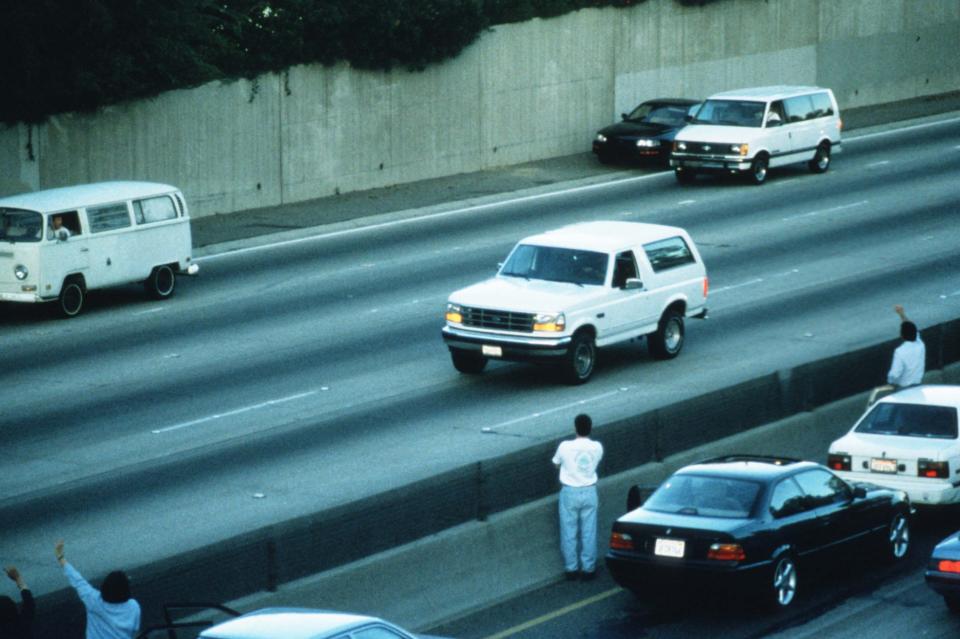 Motorists wave as police cars pursue the Ford Bronco (white, R) driven by Al Cowlings, carrying fugitive murder suspect O.J. Simpson, on a 90-minute slow-speed car chase June 17, 1994 on the 405 freeway in Los Angeles, California.