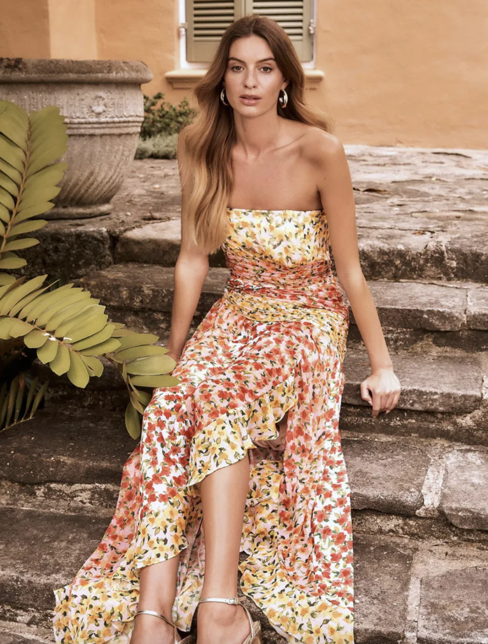 brunette model sitting on rock stairs wearing strapless orange, yellow, red and floral Rosa Printed Maxi Dress (photo via Ever New)