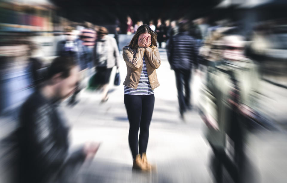 A woman with her hands covering her face stands still as people blur around her, portraying a sense of overwhelm or anxiety in a crowded place