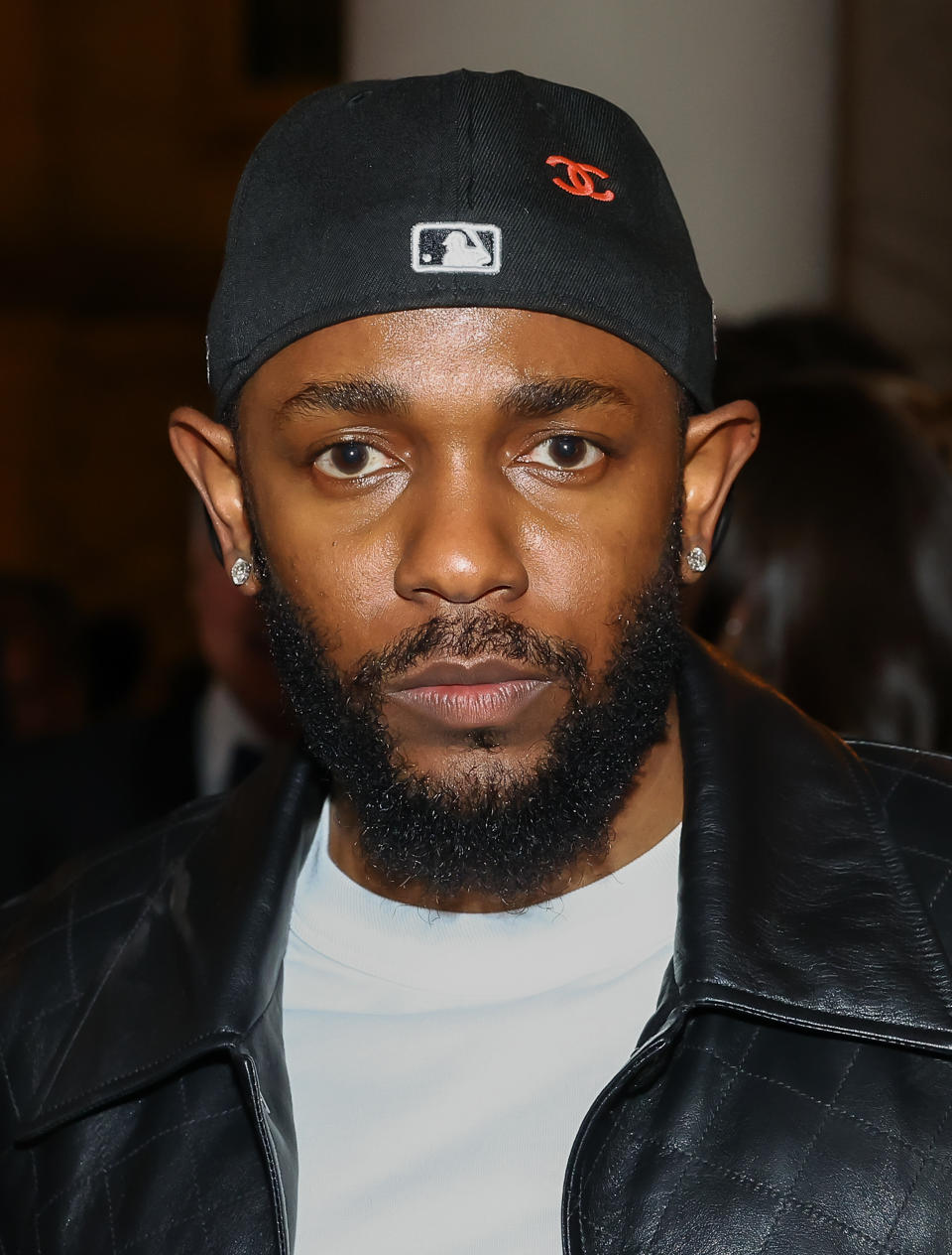 A closeup of Kendrick Lamar wearing a cap, diamond stud earrings, a shirt, and a leather jacket