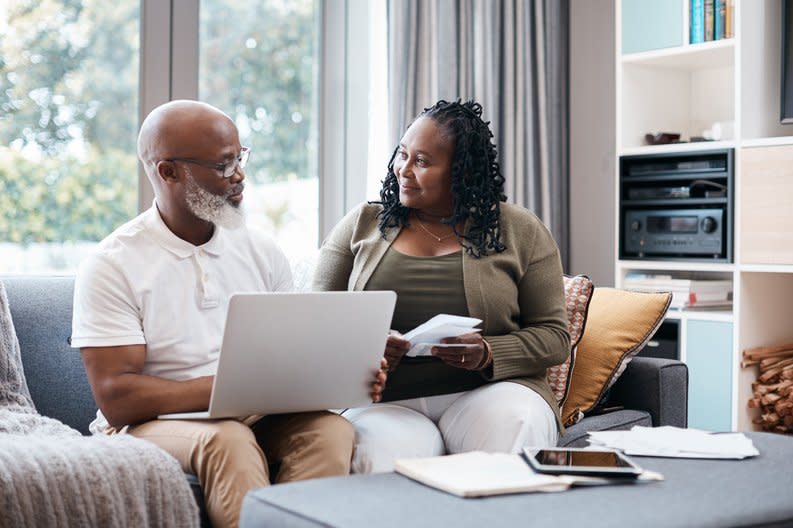 Senior couple sitting on couch together going over information on a laptop and other documents.