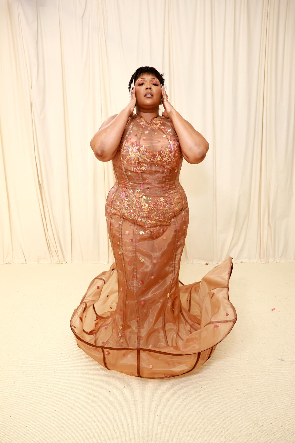 Lizzo in a floor-length, glittery gown with a trail, hands touching her hair, posing at an event