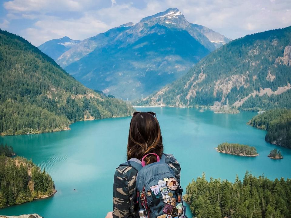 Emily, wearing a backpack covered with patches, looks out at a lake with blue waters and mountains covered in trees.