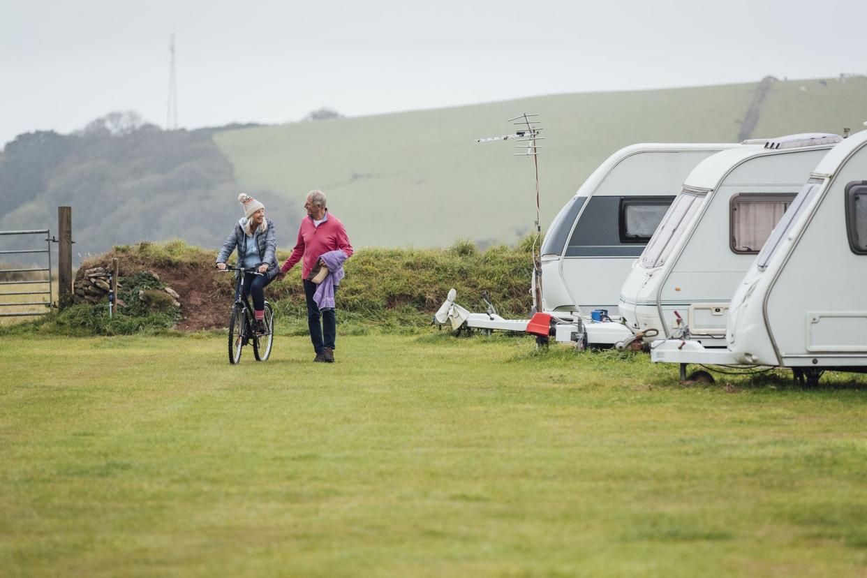 A shot of a senior woman riding a bike through a campsite in Cornwall, her husband is walking alongside her holding a towel and a wash bag.