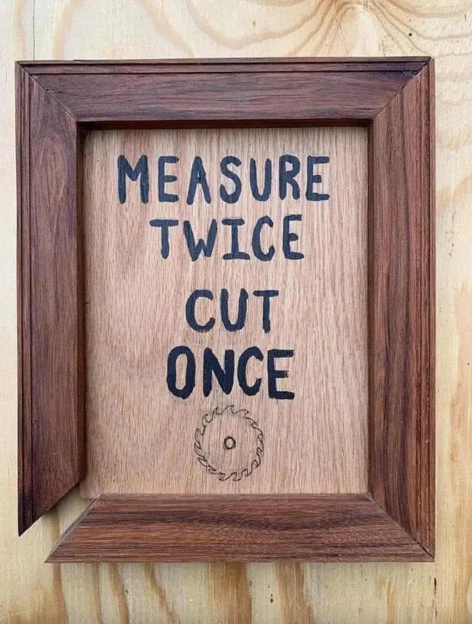 Wooden framed sign with text "MEASURE TWICE CUT ONCE" above a saw blade illustration