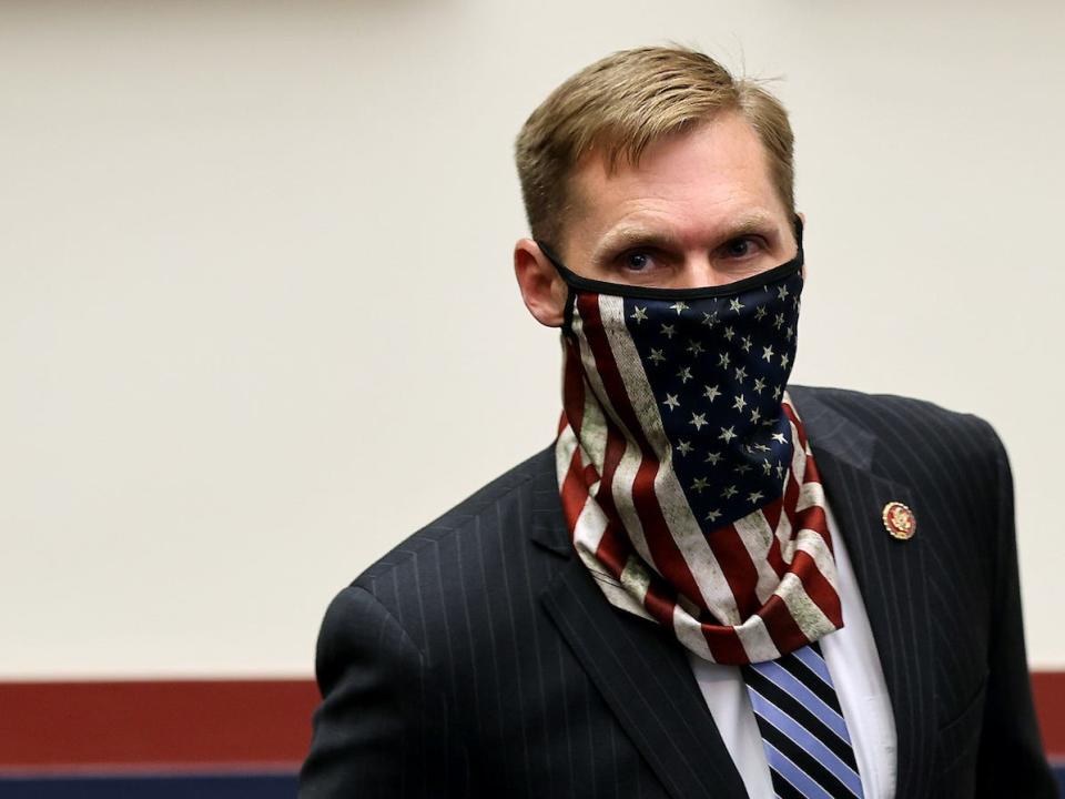 Rep. Michael Guest, sporting a mask adorned with the American flag, a dark suit, light-colored shirt, and striped tie, makes his way through a committee hearing room on Capitol Hill.