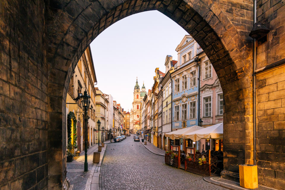 View of a European street through an archway with a clock tower in the distance and sidewalk cafes to the side