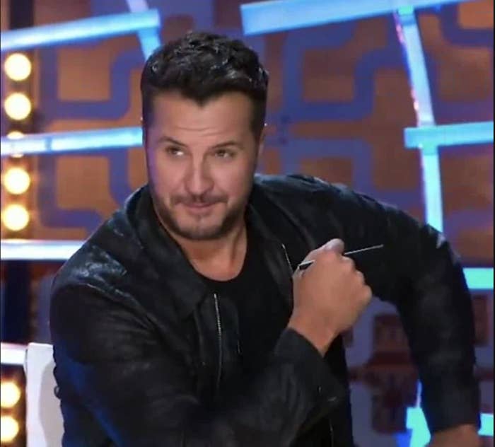 Country singer Luke Bryan sits, wearing a black jacket over a black shirt and holding what looks like a pen or pointer, with stage lights in the background