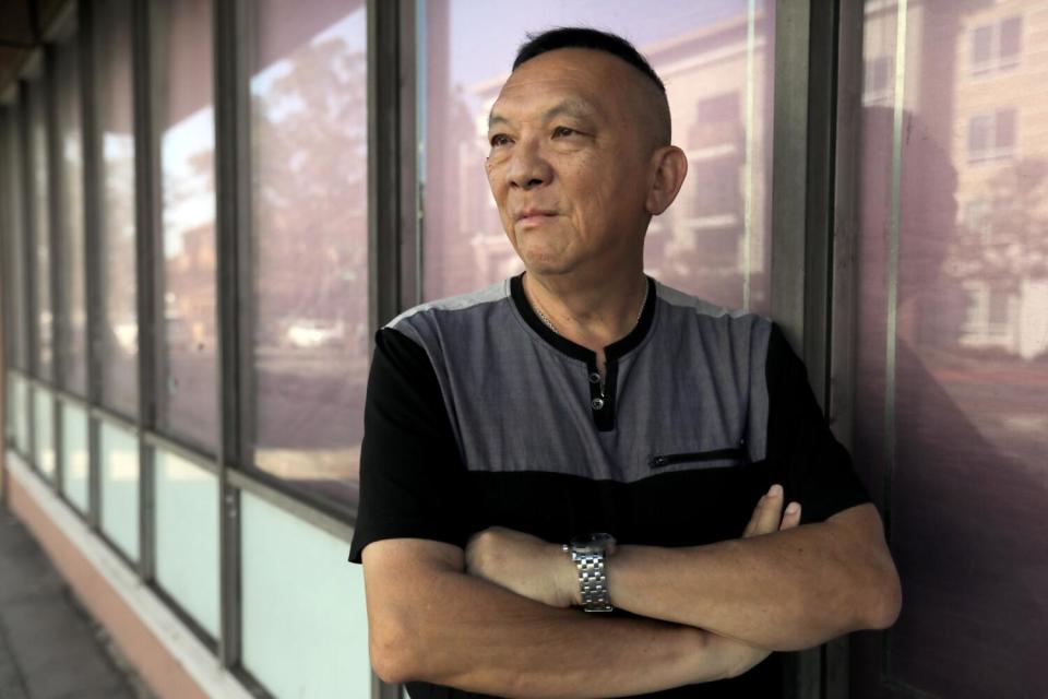 A man in a black-and-gray shirt, arms crossed, outside a building