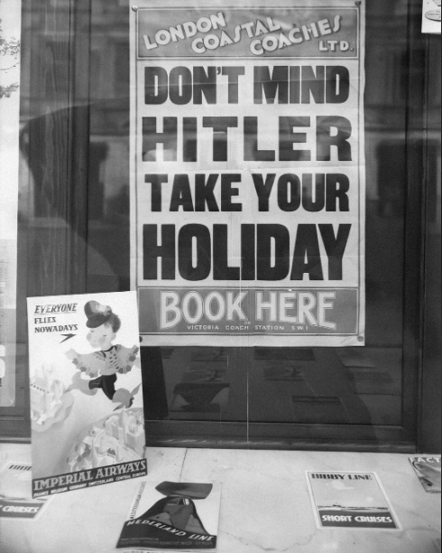 Poster in a shop window reads: "Don't mind Hitler, take your holiday. Book here, Victoria Coach Station, E.W.I". Below, Imperial Airways and other travel ads are displayed