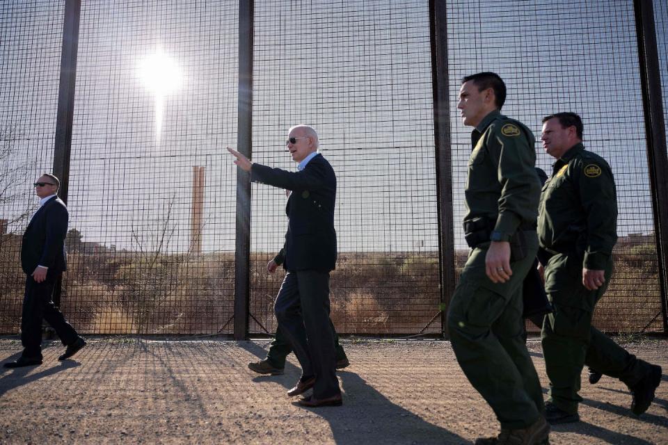 Joe Biden wears dark sunglasses and a suit and walks, in front of men in green uniforms, along a large fence. The sun shines through it.