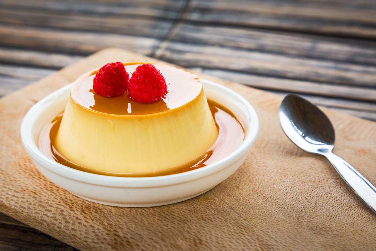 Creamy baked custard with maple syrup topped with two raspberries in a white dessert plate on a wooden cutting board with a dessert spoon with a blurred wooden table background