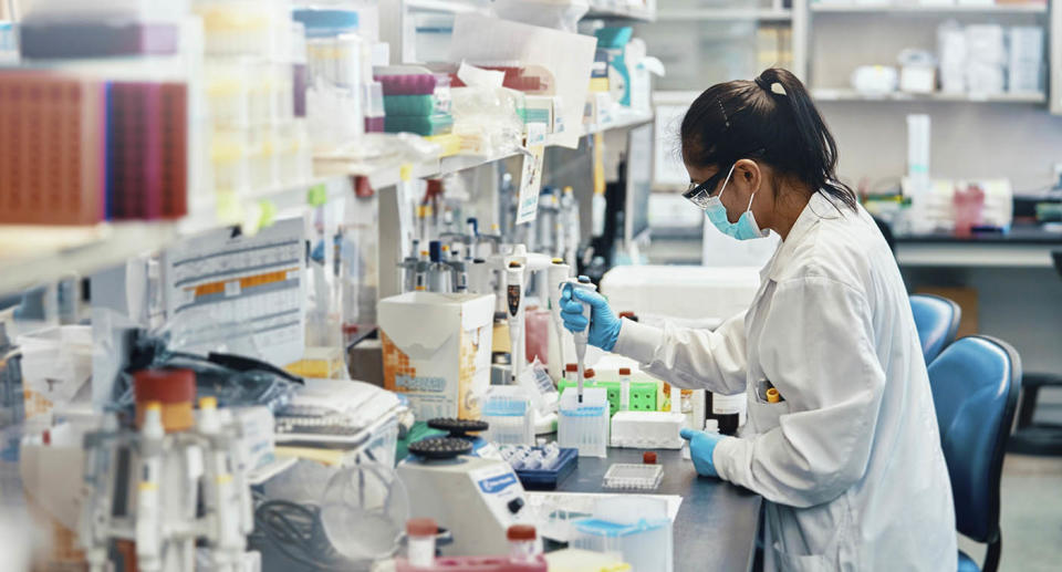 Female scientist in a lab testing something sitting down wearing white lab coat and safety goggles. 