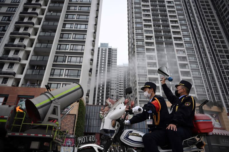 Man holding a loudspeaker sits on a motorcycle as it travels past a sanitizing vehicle disinfecting the public space near residential buildings in Guangzhou