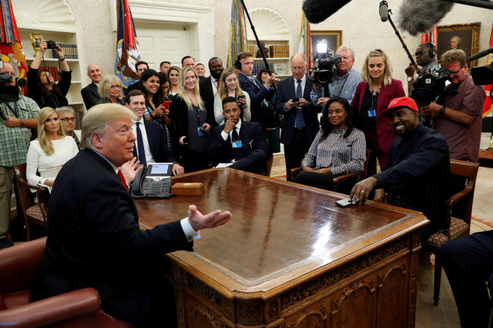 Donald Trump, behind his desk in the Oval Office, with a throng of cameramen and visitors around him, makes a quip that most of the assembled company finds highly amusing. Seated are Ivanka Trump and Jared Kushner, as well as Kanye West, wearing a red MAGA baseball cap.