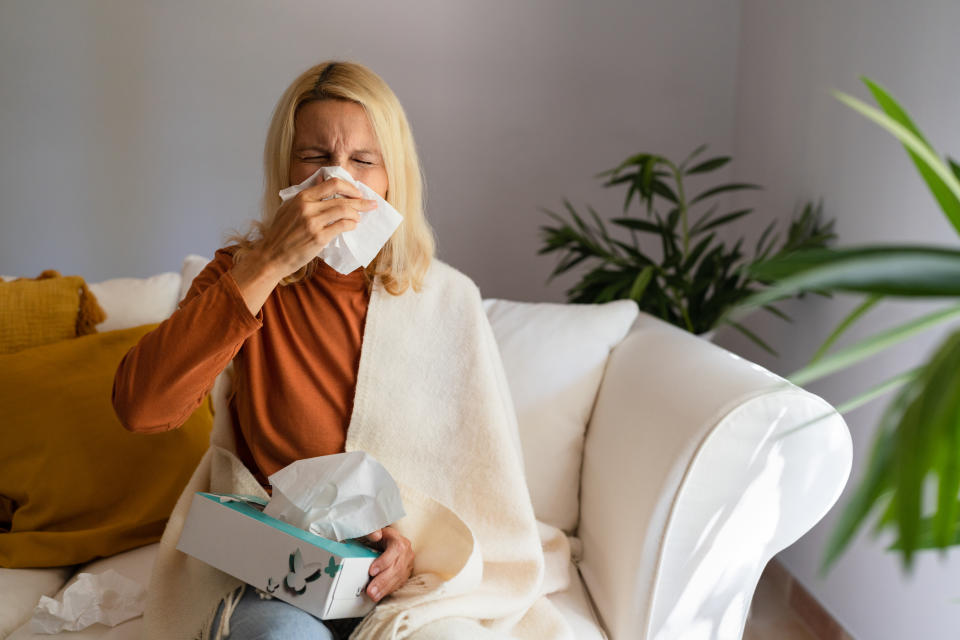 Sickwoman wrapped in blanket sitting on sofa and blowing her nose with a tissue. Preventing infection is crucial when it comes to flu season. (Getty)