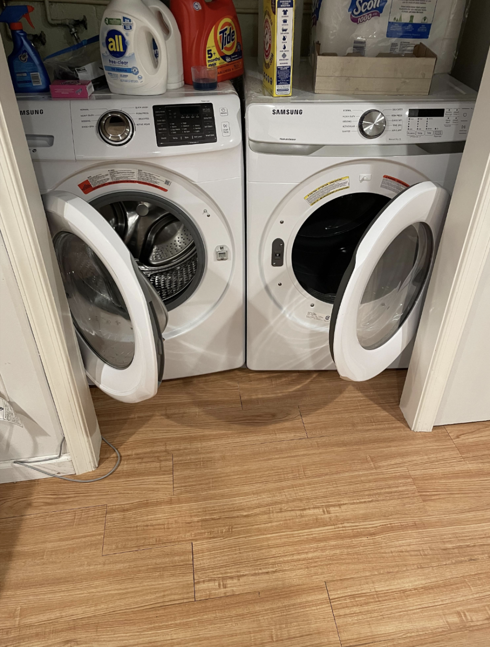 Two open Samsung front-loading laundry machines in a closet with various detergent and cleaning supplies on top