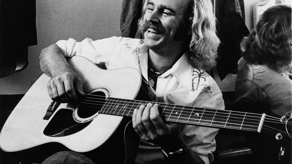 jimmy buffett smiles while playing a guitar in this black and white photo, he wears a hawaiian t shirt and sits back against a mirror