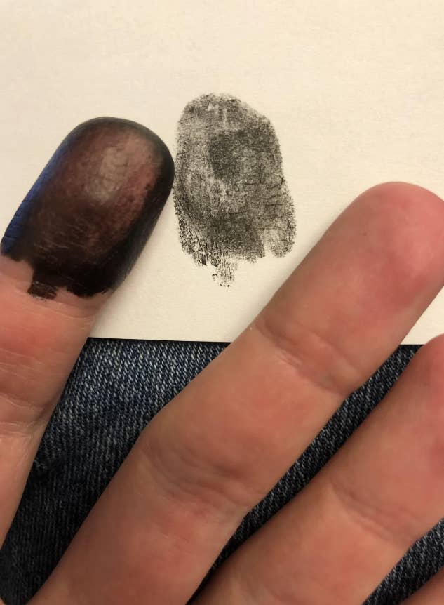 A thumb with ink on it and an ink thumbprint on a white surface beside it. Other fingers resting on a denim fabric