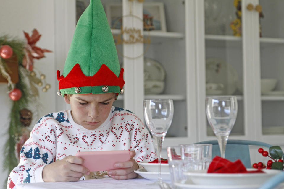 A young boy wearing an elf hat holds a smartphone with both his hands while at a decorated table, with his brow furrowed and a serious expression on his face