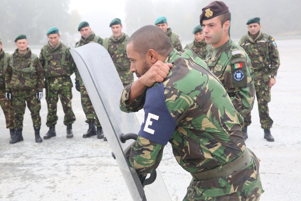 A photo of a man in army fatigues, holding a riot shield, with his arm across his chest