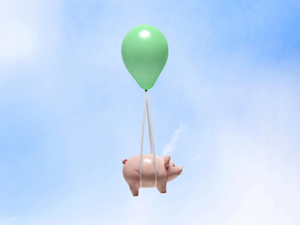 Green helium balloon carrying pink piggy bank with white strings on blue sky.