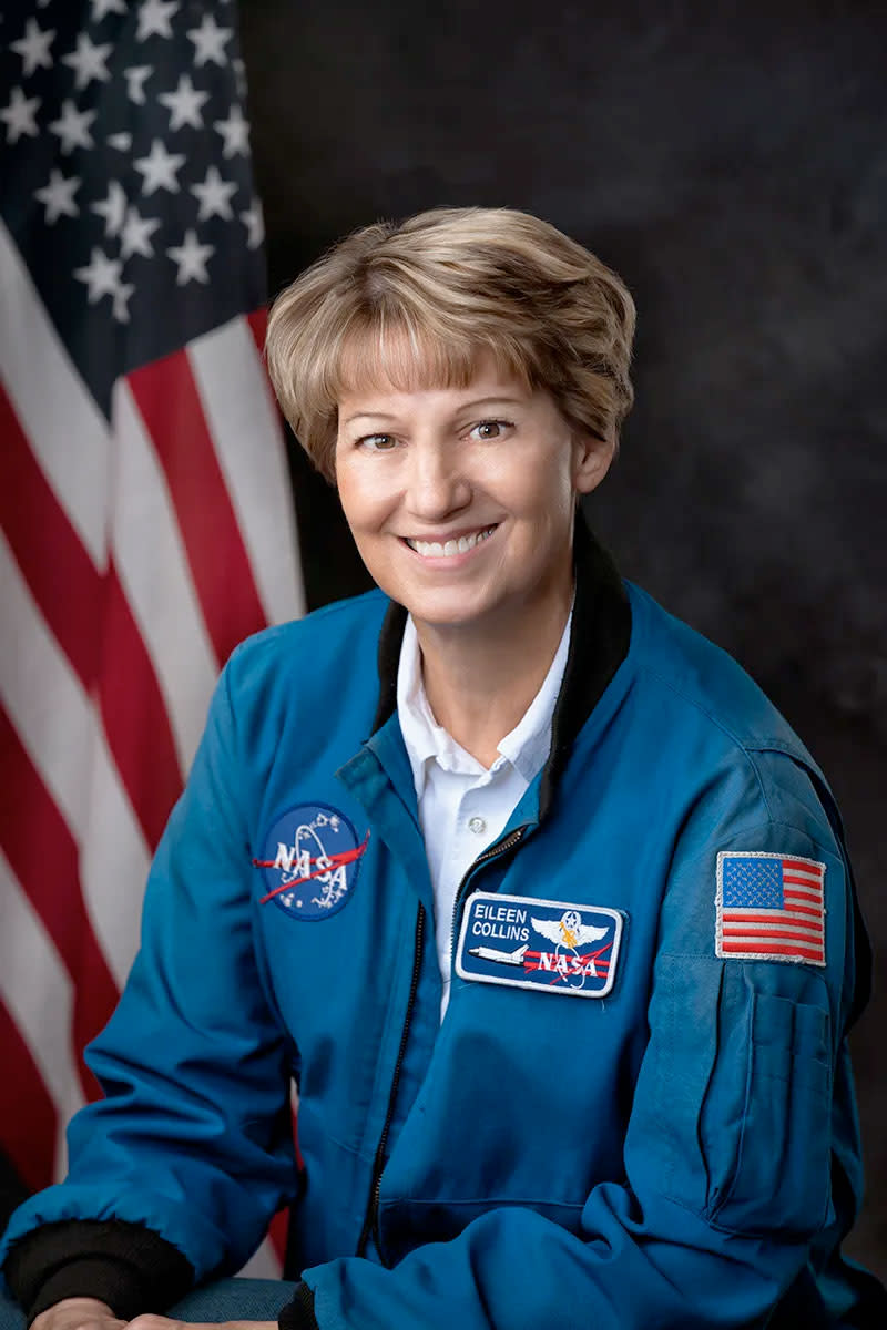 a smiling woman in a blue flight suit sitting in front of an American flag
