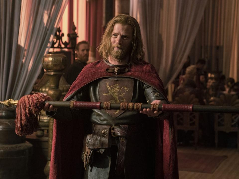 A red-haired man wearing a red cape presents a gold spear.