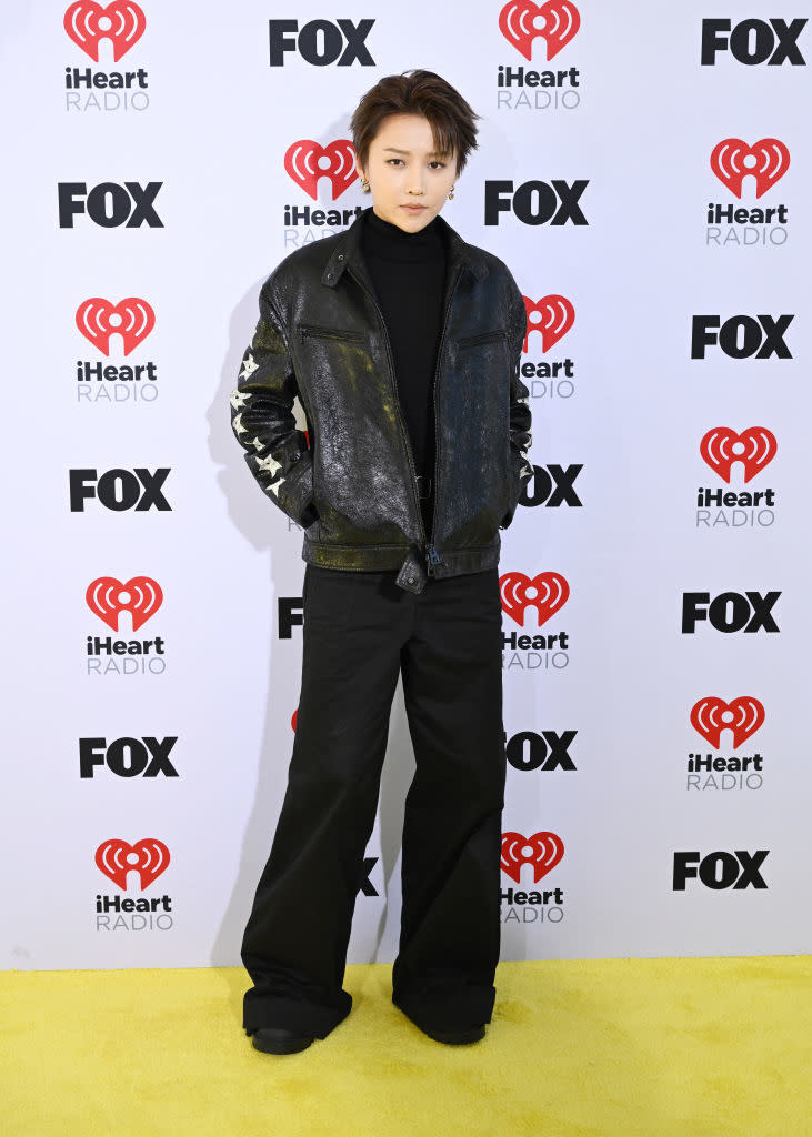 Person standing on yellow carpet, iHeartRadio backdrop, in black turtleneck & patterned jacket