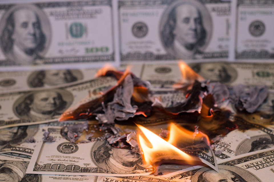 A small pile of hundred dollar bills on fire, with other hundred dollar bills lined up in the background.