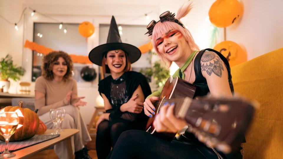 three young women in costumes enjoying fun at halloween party activities, singing, playing guitar