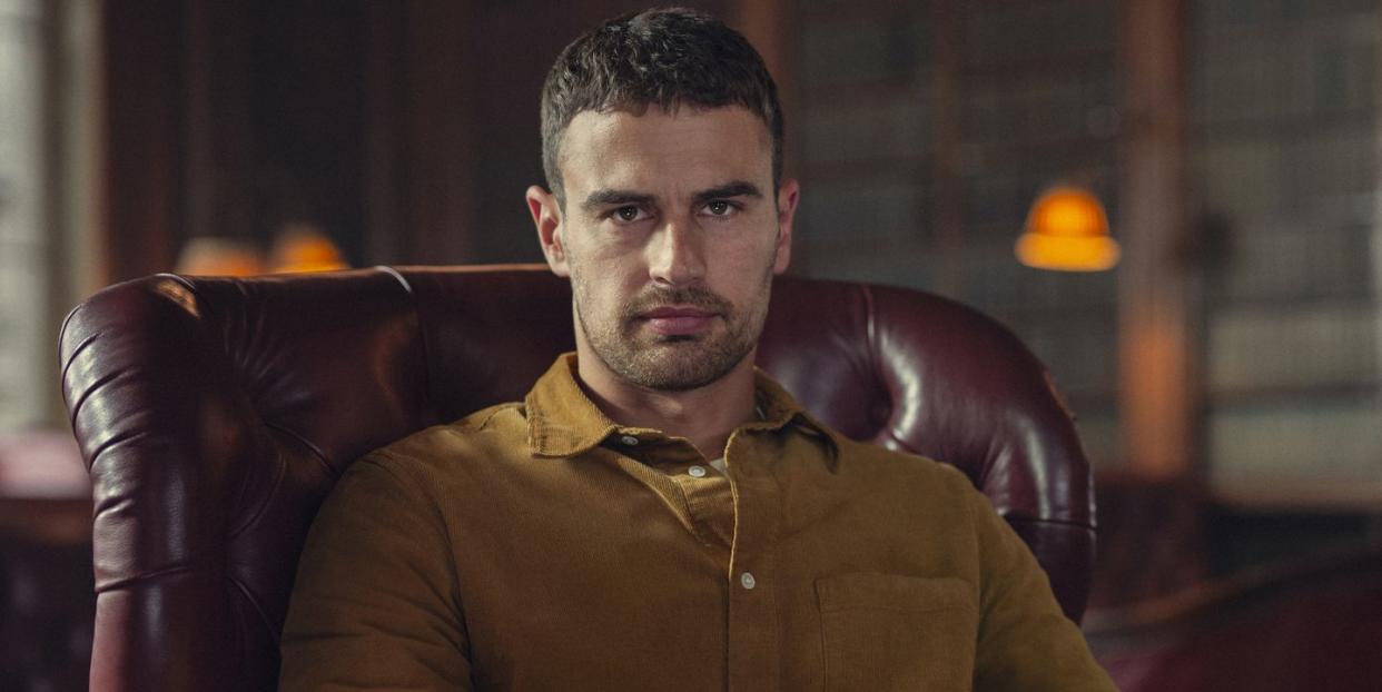 theo james in the gentlemen, a young man sits in an armchair looking at the camera and holding a wine glass